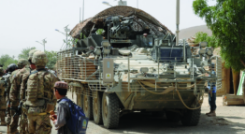 The rear view of a Stryker armored vehicle with camouflage netting, on patrol in a village street with soldiers and a local child nearby.